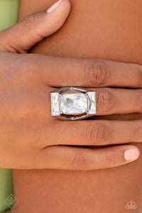 An exaggerated, oversized, rectangular frame encases a large, white, emerald-cut gem, showcasing its scintillating shimmer. Skinny white gems chiseled into rectangular bars are pressed into the top and bottom of the industrial frame, resulting in an unapologetic statement piece with unmatched sparkle. Features a stretchy band for a flexible fit.